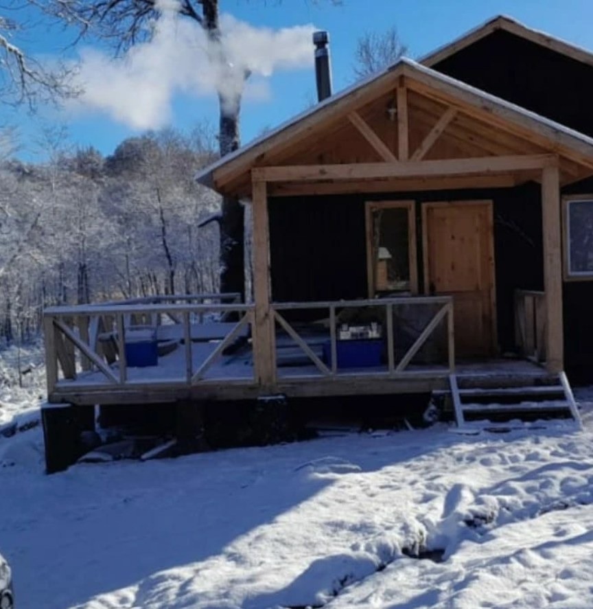 Cabaña de madera negra con nieve en la terraza y humo saliendo de la chimenea