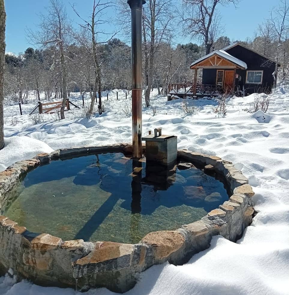Tinaja de piedra con agua transparente rodeada de nieve, con una cabaña al fondo