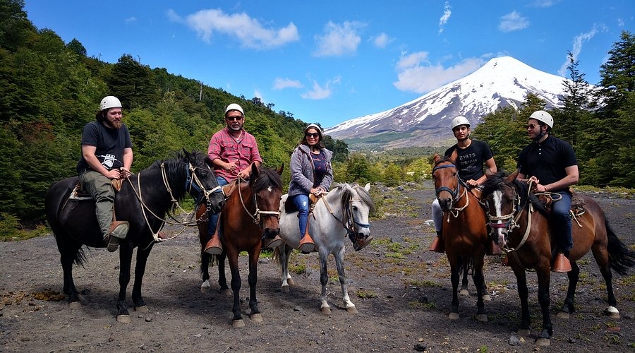 Grupo de jinetes con cascos en un sendero de montaña, con un volcán nevado al fondo